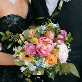 Colorful hand-tied bouquet of pink peonies, orange ranunculus, and mixed blooms held by a dressed-up couple