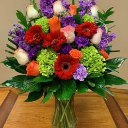 Mixed bouquet of red gerbera daisies, orange and white roses, and purple flowers in a glass vase