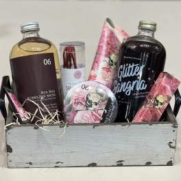 Wooden crate gift set with bath soak, sangria mixer, and pink floral skull-themed self-care items.