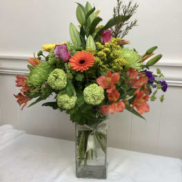 Tall mixed bouquet of orange, green, yellow and pink flowers in a clear rectangular glass vase with ribbon