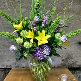 Tall bouquet of yellow lilies, purple blooms, and green flowers in a glass vase
