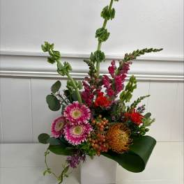 Tall modern arrangement with pink gerberas, snapdragons, orange protea and roses in a white cube vase