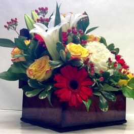 Mixed flower arrangement in a dark square box with lilies, roses, and gerbera daisies