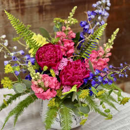 Low arrangement of bright pink and blue flowers with ferns in a white patterned ceramic container