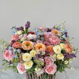 Lush arrangement of pink, peach and yellow roses with lilac and blue blooms in a rustic twig container