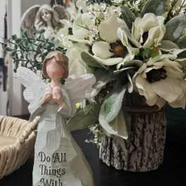 White floral arrangement with an angel figurine and a carved "Do All Things With Love" statue