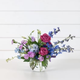 Mixed bouquet of pink roses, blue delphinium, and purple blooms in a glass vase