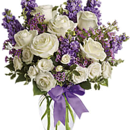 White roses and purple flowers arranged in a clear glass vase with a purple ribbon