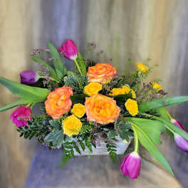 Bouquet of orange and yellow roses with purple and pink tulips in a white container