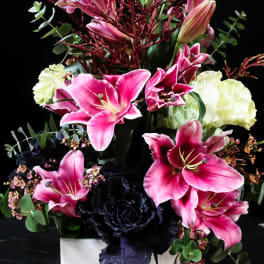 Pink lilies and dark purple flowers in a white square vase