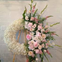 Pink and white floral funeral wreath on an easel beside a tall mixed arrangement