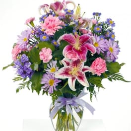 Pink lilies, carnations, and purple daisies in a clear glass vase with a lavender ribbon