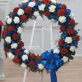 Patriotic wreath of red, white, and blue flowers on a white easel with a large blue bow.
