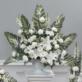 Tall white arrangement with lilies, roses, carnations, and daisies in a white urn with palm leaves