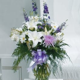 Tall vase arrangement with white lilies, daisies, lavender mums, and purple blooms with a lavender bow.