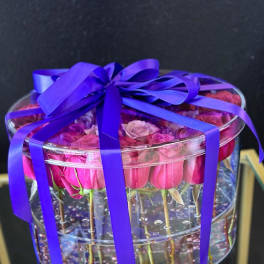 Pink roses in a clear round box tied with a purple ribbon