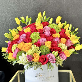 Bright arrangement of multicolor roses, yellow tulips, lilies, and green blooms in a white hat box.