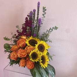 Bouquet of orange roses, sunflowers, and purple flowers in a clear vase