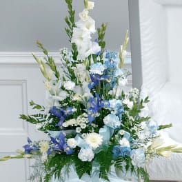 Tall white and blue sympathy flower arrangement in a white pedestal vase beside an open casket