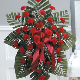 Standing spray of red carnations with baby’s breath and large palm leaves on an easel.