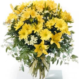 Tall arrangement of yellow lilies and daisies with white accents in a clear glass vase with ribbon