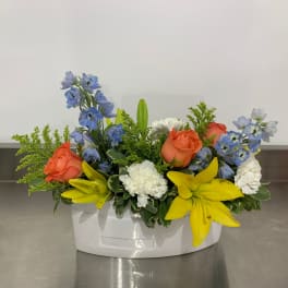 Mixed floral arrangement in a white container with orange roses, blue flowers, and yellow lilies