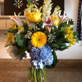 Mixed bouquet with yellow lilies, blue hydrangeas, and a yellow gerbera daisy in a glass vase