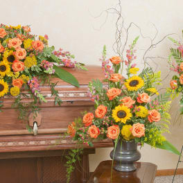 Three floral funeral arrangements with orange roses and sunflowers around a casket.