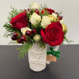 Compact arrangement of red and white roses with berries and evergreens in a white Santa mug.