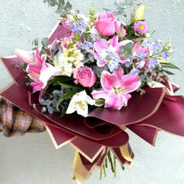 Hand-tied bouquet of pink roses, lilies, and mixed blooms wrapped in burgundy and gold paper.