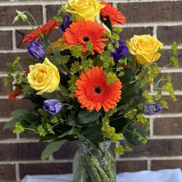 Bouquet of yellow roses, orange gerbera daisies, and purple blooms in a glass vase