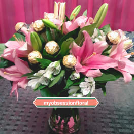 Pink lilies and white flowers arranged in a glass vase with gold-wrapped chocolates