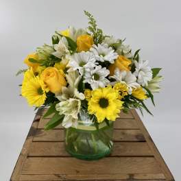 Yellow roses and white daisies in a green glass vase on a wooden crate
