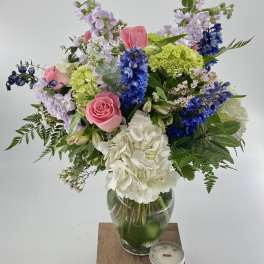 Mixed bouquet of pink roses, blue delphinium, and white hydrangeas in a glass vase with a small candle.