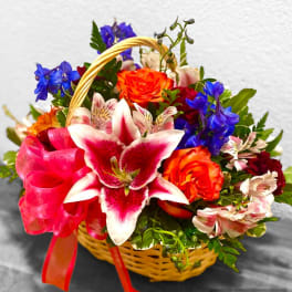 Basket arrangement with pink lilies, orange roses, blue flowers, and a pink ribbon