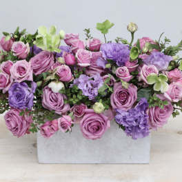 Low rectangular arrangement of pink and lavender roses with purple blooms in a white container