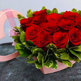 Red roses arranged in a pink heart-shaped box with green foliage