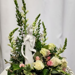 White angel figurine surrounded by cream and pink roses in a floral arrangement