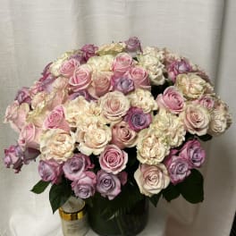 Large arrangement of pastel pink, cream, and lavender roses in a glass vase with a candle beside it