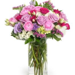 Tall glass vase of pink and lavender roses with mixed pastel flowers in a rounded arrangement