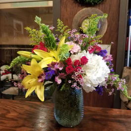 Mixed bouquet with yellow lilies, red roses, white hydrangea, and purple accents in a glass vase