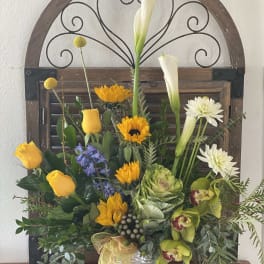 Mixed floral arrangement with yellow roses, white calla lilies, and sunflowers in a silver vase