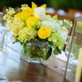 Yellow lilies and roses with white blooms in a clear cube vase on a table