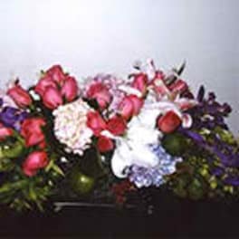 Low, rectangular arrangement of pink roses, white lilies, and mixed blooms in a dark container