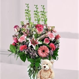 Pink mixed flower arrangement with lilies, roses, and gerbera daisies in a glass vase with a tan teddy bear.