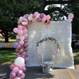 Pink and white balloon arch beside a white floral wall with a chair in front