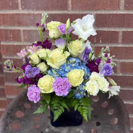 Bouquet of yellow roses, blue hydrangea, and purple flowers in a dark vase