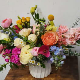 Low arrangement of mixed pastel roses, ranunculus, calla lily, and other blooms in a white vase.