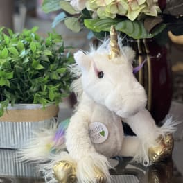 Plush unicorn with gold horn and hooves beside potted greenery and hydrangeas