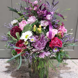 Tall mixed bouquet of pink roses, white daisies, and purple blooms in a clear glass vase with a purple ribbon.